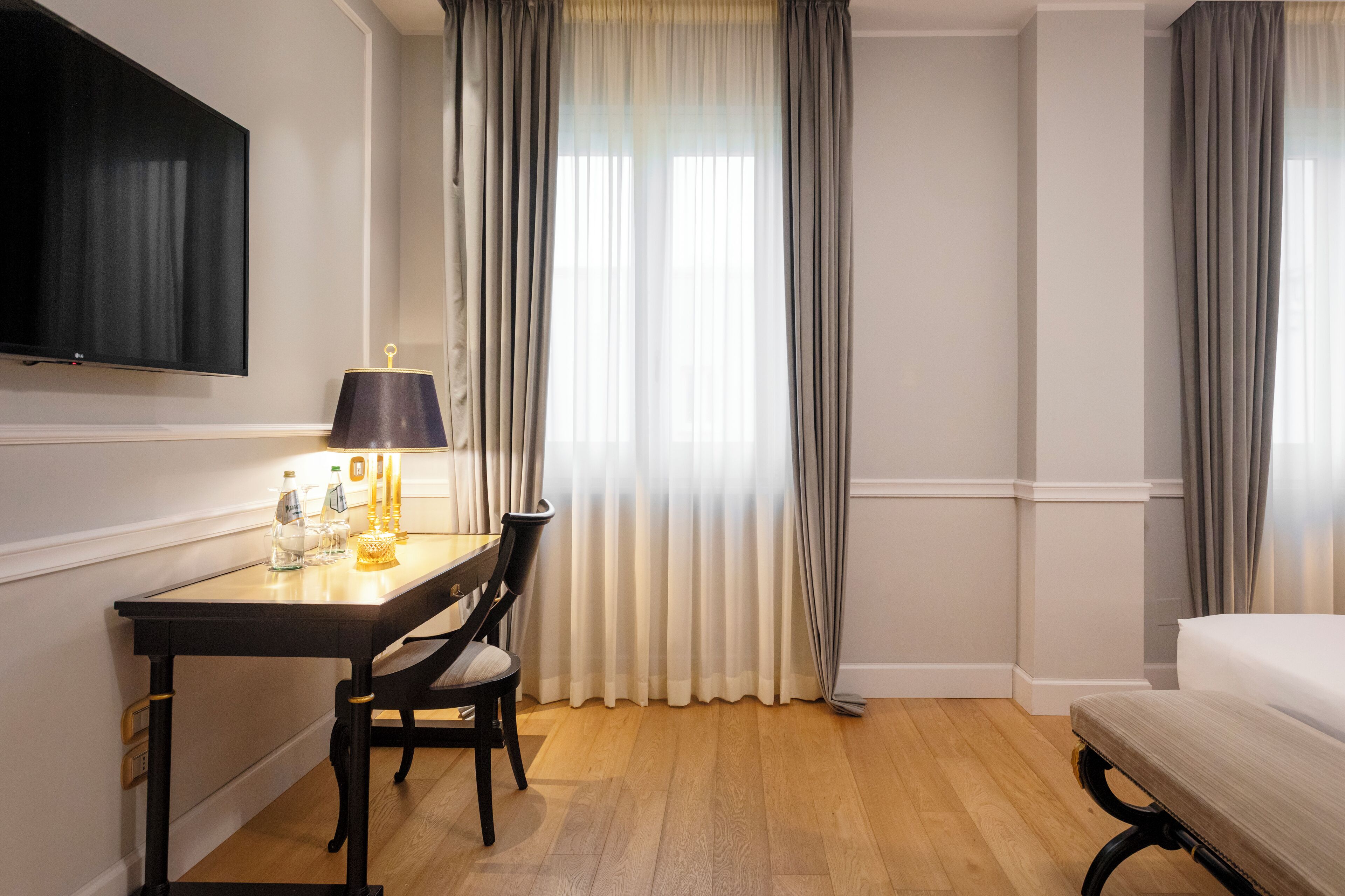 A hotel room with a bed, a desk with a lamp, a chair, and a TV mounted on the wall.