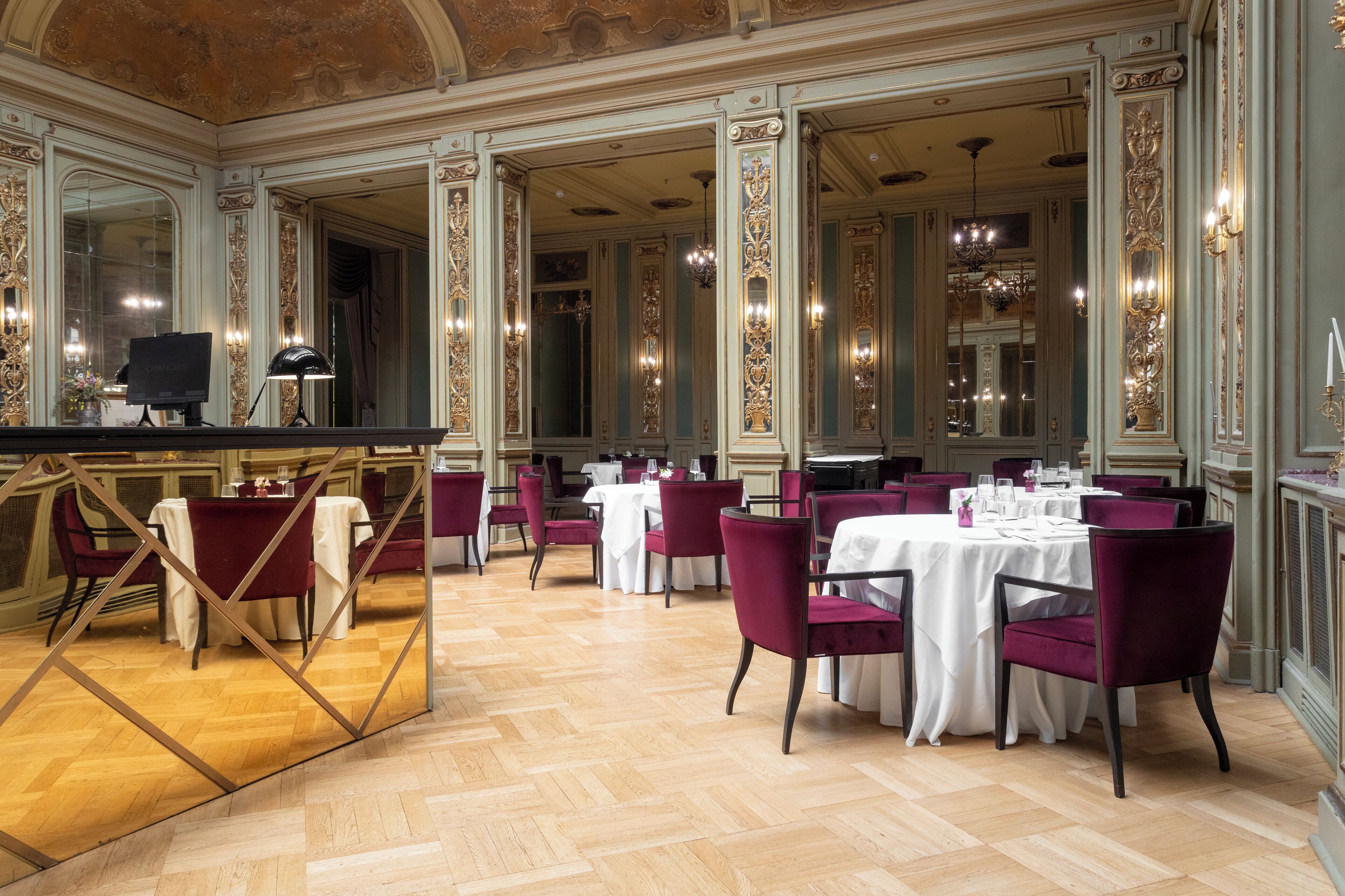 A spacious dining area with high ceilings, ornate columns, and chandeliers. Tables are set with white tablecloths and red chairs.
