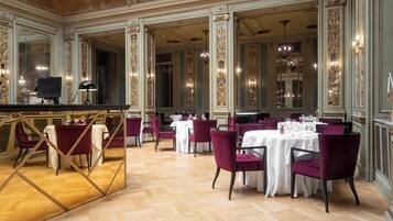 A spacious dining area with high ceilings, ornate columns, and chandeliers. Tables are set with white tablecloths and red chairs.