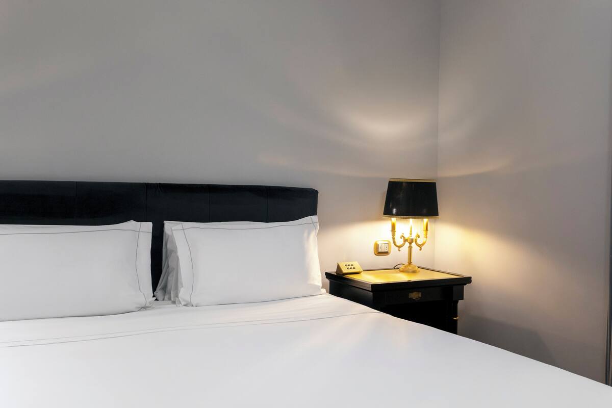 A neatly made bed with white linens and a black headboard, a bedside table with a lamp, and a wall-mounted telephone.