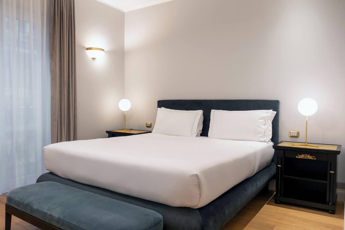 A neatly made bed with white linens, two bedside tables with lamps, a bench, and a wall-mounted light fixture.