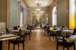 Un comedor largo y elegante con techos altos, molduras ornamentales y un candelabro. Las mesas están dispuestas con vajilla fina, cristalería y sillas tapizadas en rojo.