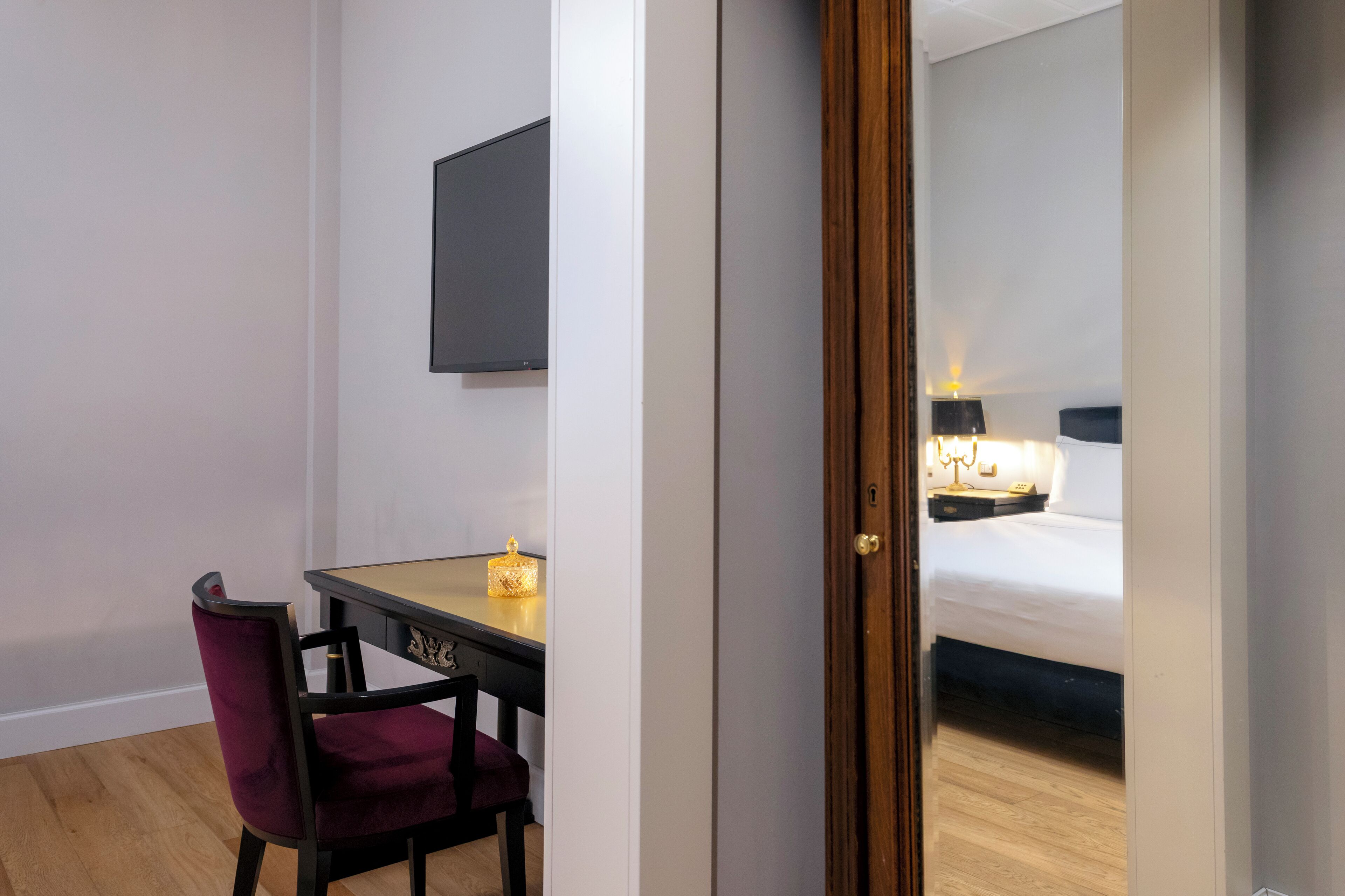 A hotel room with a wooden floor, a desk with a chair, a bed, a TV mounted on the wall, and a nightstand with a lamp.