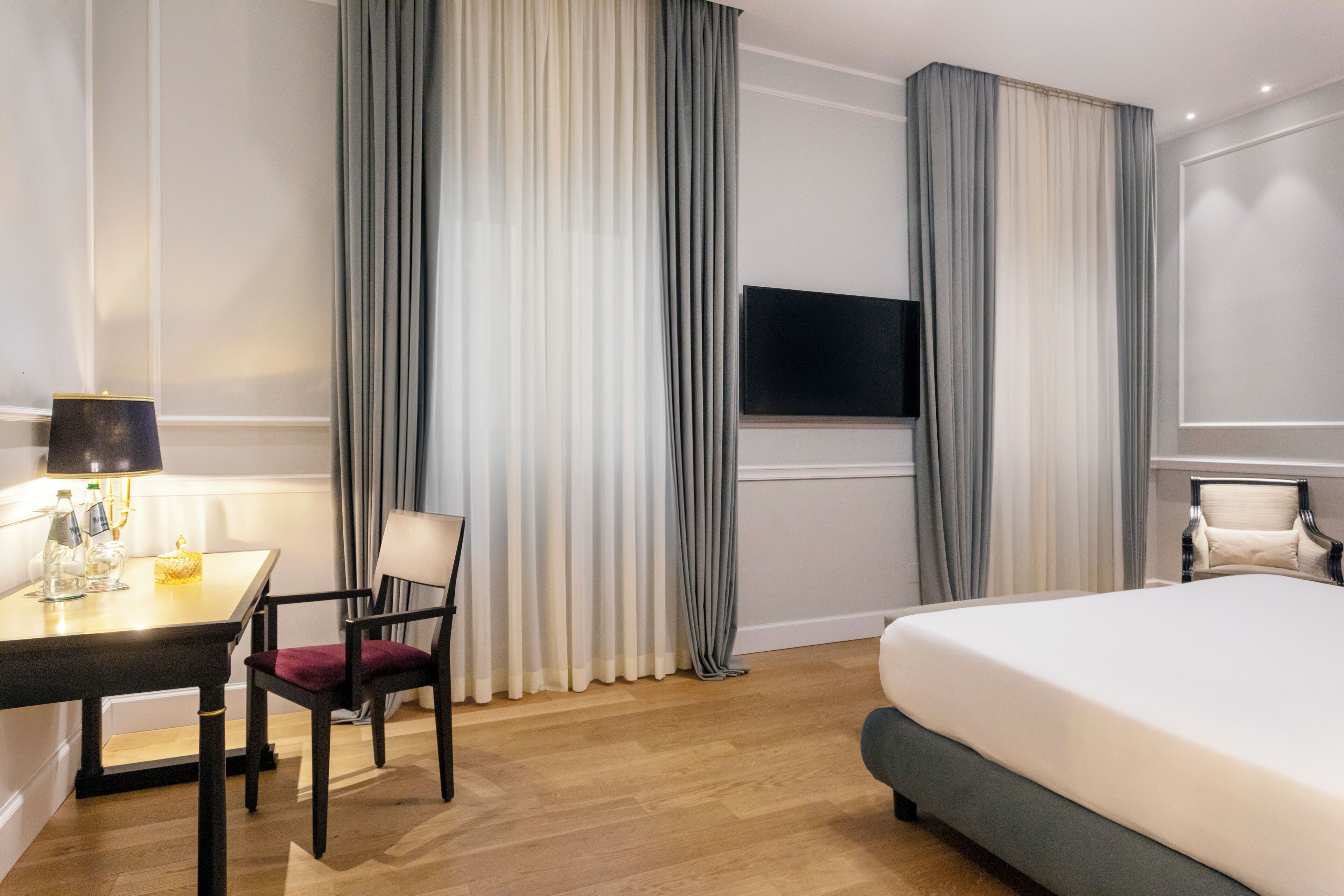 Een hotelkamer met een bed, een bureau met een lamp, een stoel, een televisie en gordijnen.