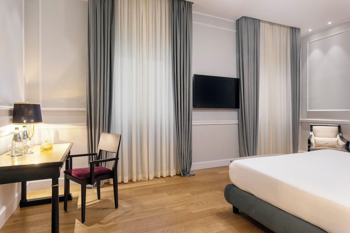 A hotel room with a bed, a desk with a lamp, a chair, a TV, and curtains.