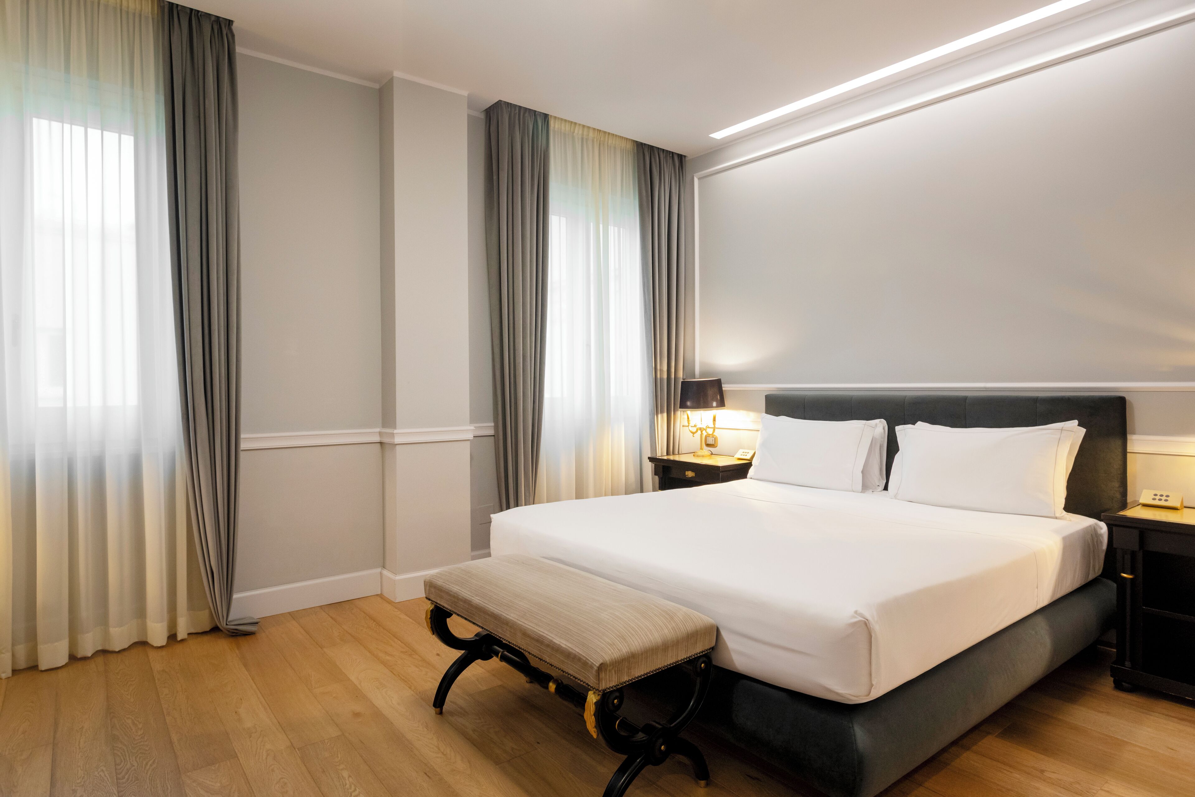 A hotel room with a large bed, a bench, a nightstand with a lamp, and curtains.