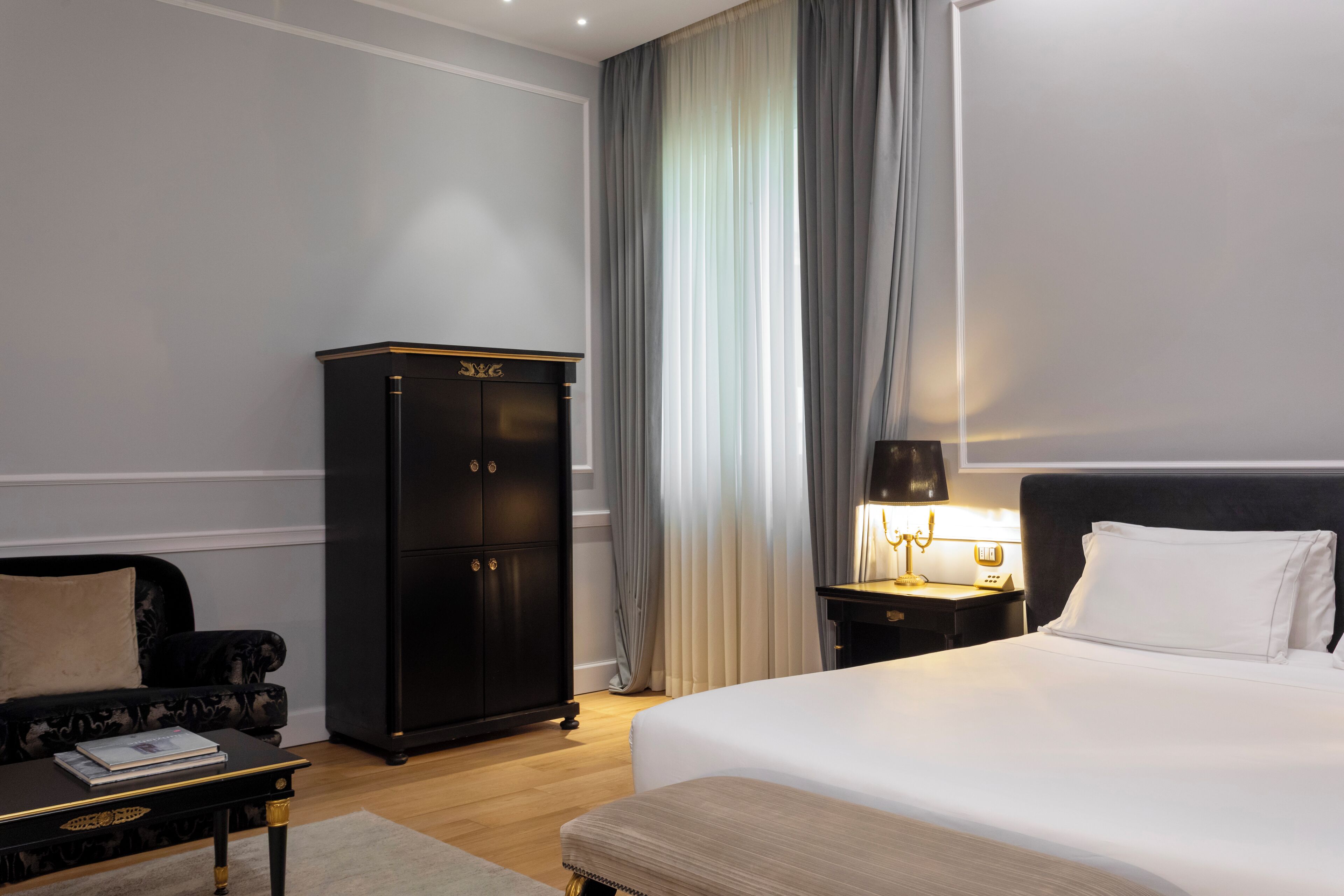 A hotel room with a large bed, a dark wooden wardrobe, a small table with a lamp, and a sofa.