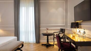 A hotel room with a bed, a desk with a chair, a small table with a lamp, and a TV mounted on the wall.