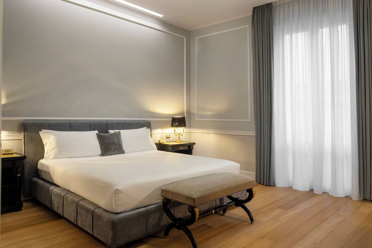 A neatly made bed with white linens and a grey headboard, flanked by bedside tables with lamps. A bench is placed in front of the bed.