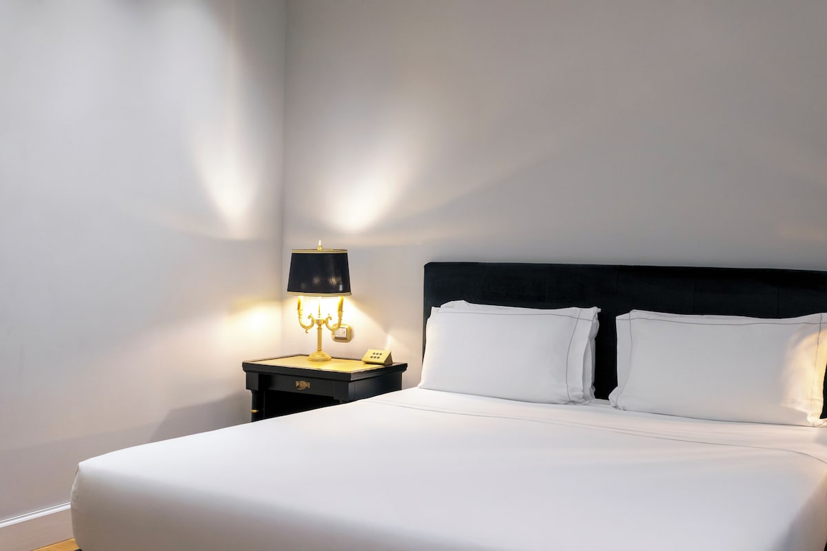 A neatly made bed with white linens and two pillows, a black headboard, and a bedside table with a lamp.