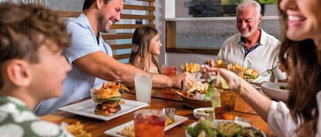 Una familia cenando en un restaurante con una mesa de madera, varios platos y un letrero al fondo.