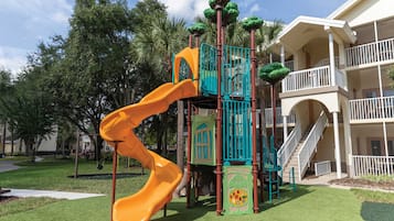 A playground with a slide and climbing structures, surrounded by a well-maintained lawn and palm trees.