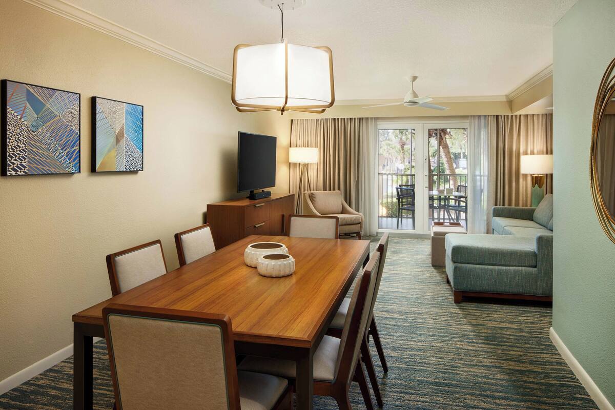 A hotel room with a wooden dining table, chairs, a television, a sofa, and a balcony with curtains.