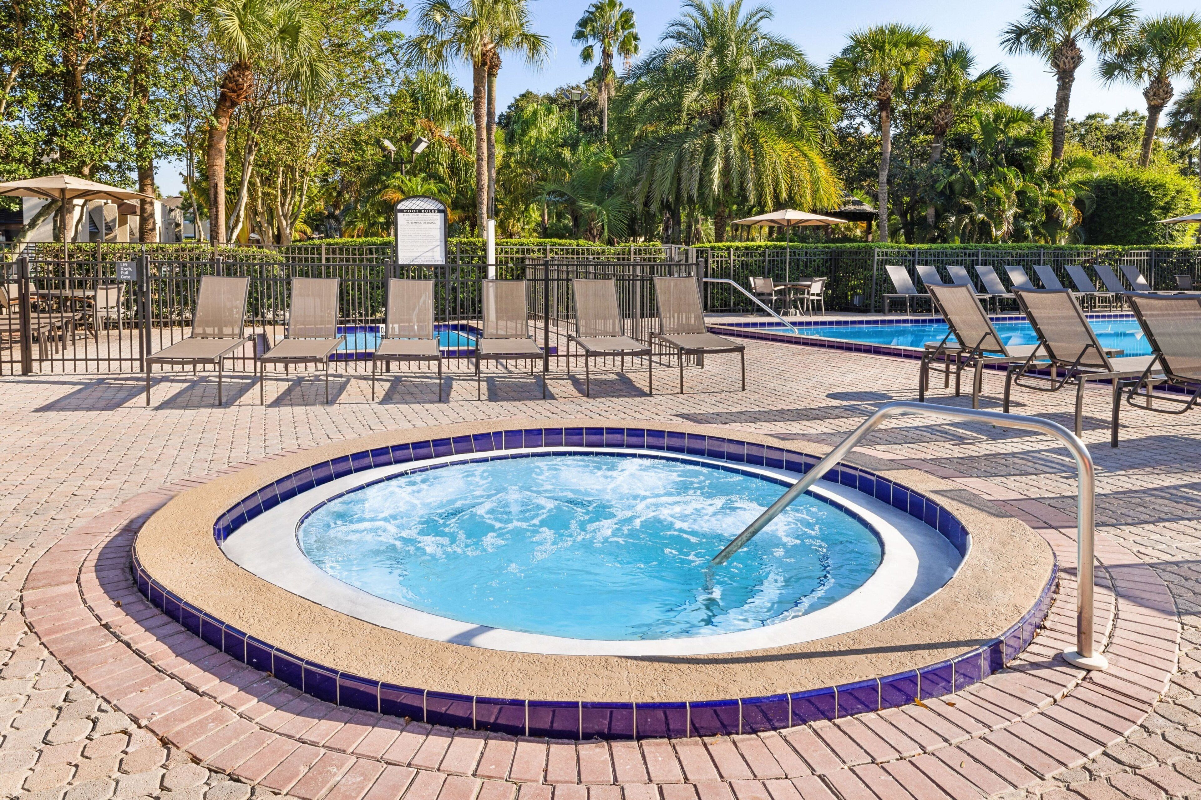 A hot tub surrounded by lounge chairs and a pool area, with palm trees and a fence in the background.
