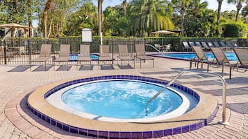 A hot tub surrounded by lounge chairs and a pool area, with palm trees and a fence in the background.
