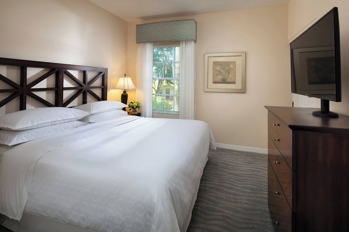 A neatly made bed with white linens, a wooden headboard, a bedside lamp, a television on a wooden dresser, and a window with a view of trees.