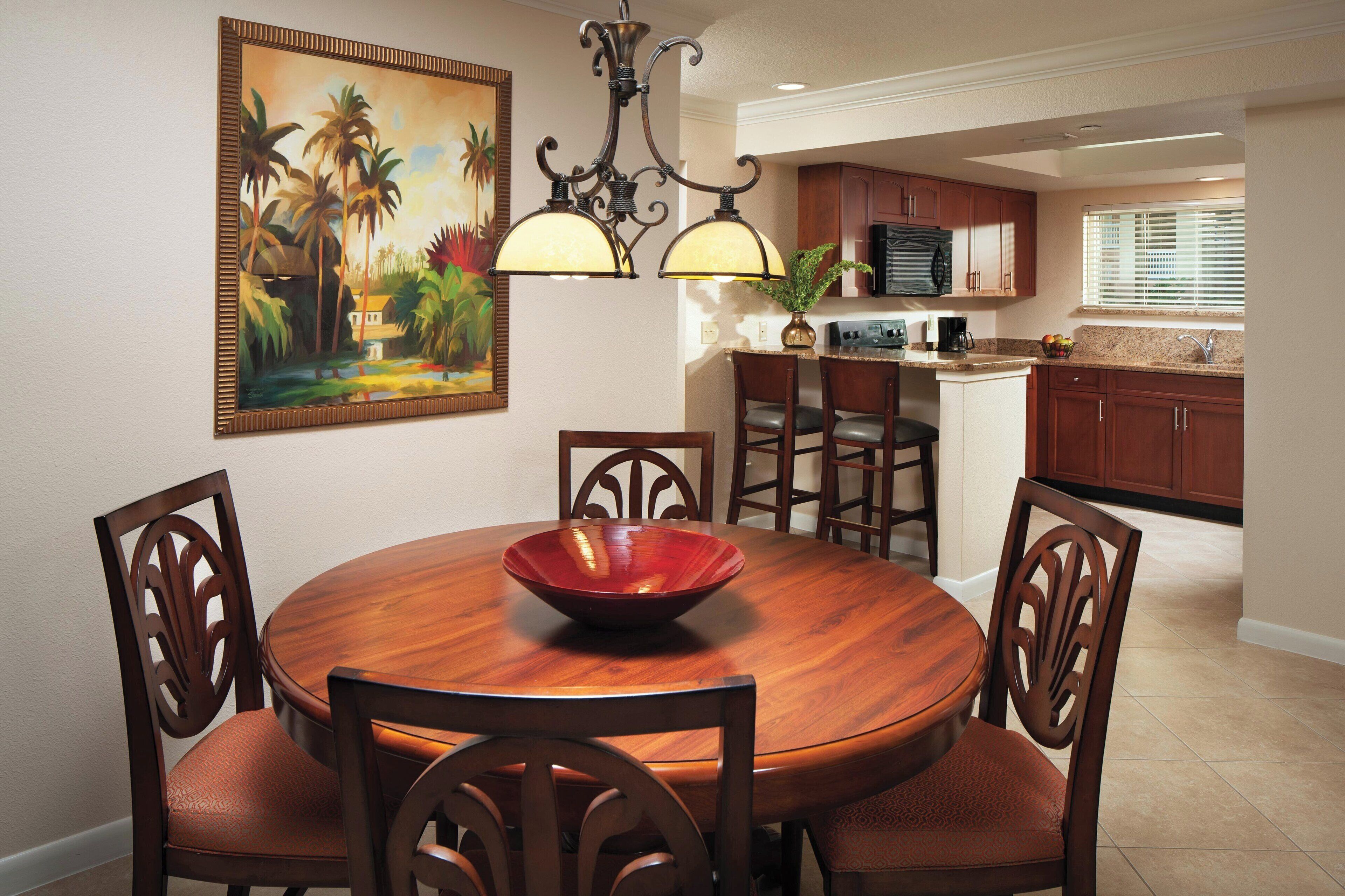 A dining area with a round wooden table, four chairs, and a red bowl. There is a painting on the wall and a kitchen in the background.
