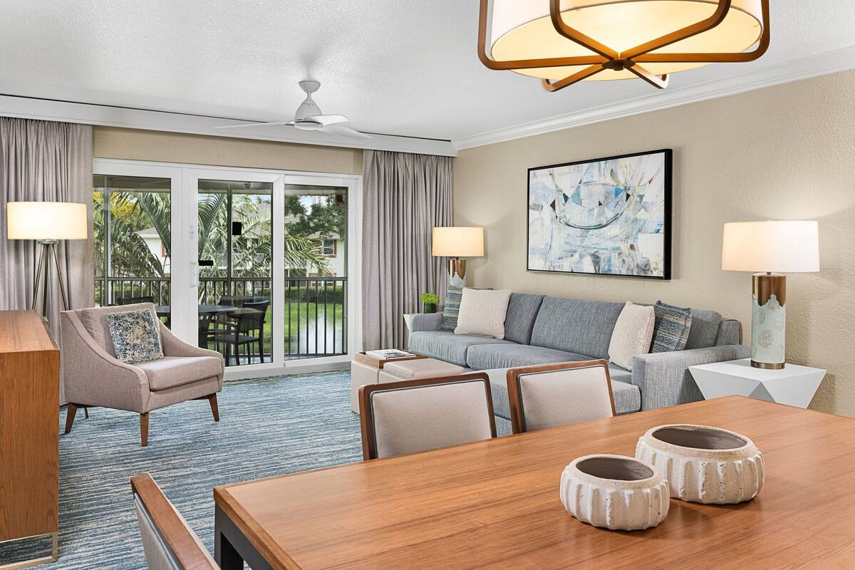 A modern hotel room with a dining area, a sofa, a chair, and a large window with curtains.