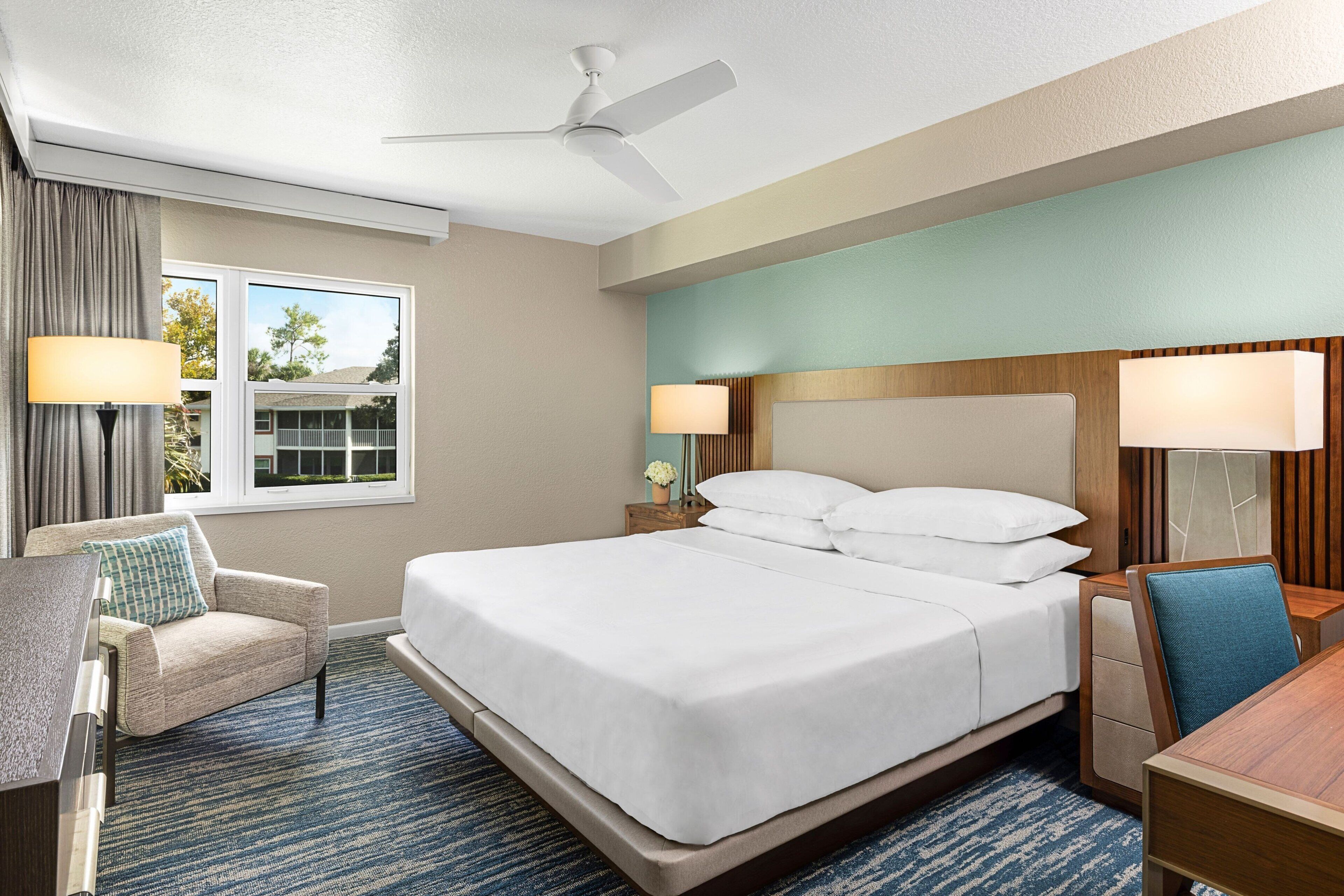 Ein Hotelzimmer mit einem großen Bett, einem Schreibtisch, einem Stuhl und einem fensterbepflanzten Fenster.