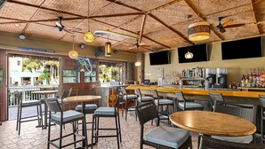 Un espace bar aménagé avec des meubles en osier, des ventilateurs de plafond et un comptoir où sont exposées différentes bouteilles.