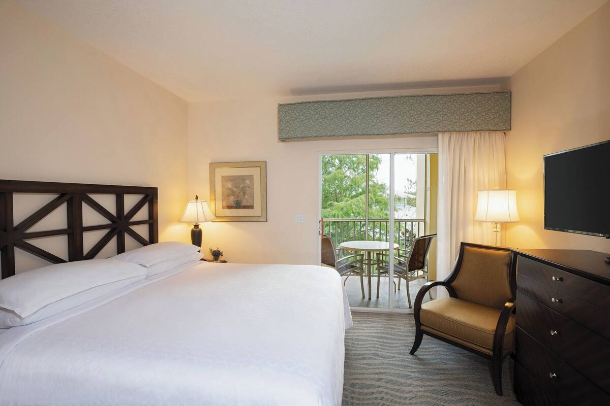 A hotel room with a large bed, a desk, a chair, a television, and a balcony with a table and chairs.