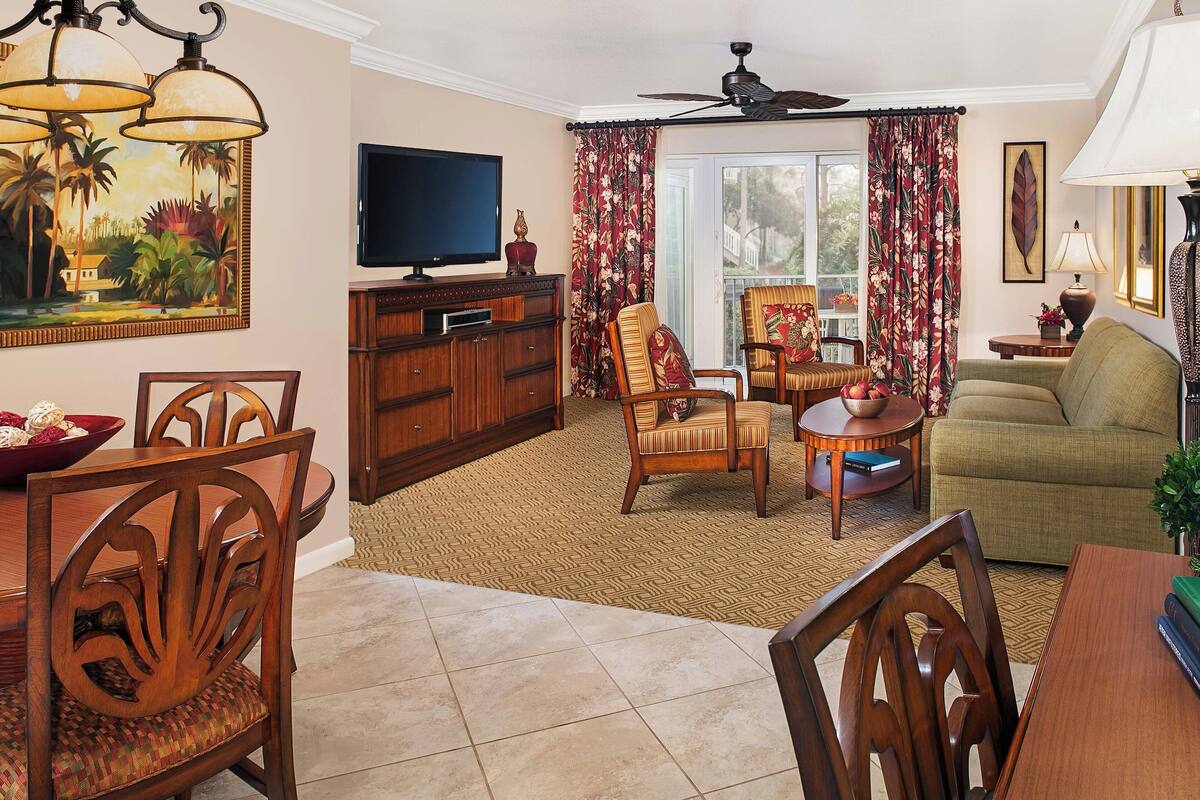 A living room with a dining area, a television, a ceiling fan, and a large window with curtains.
