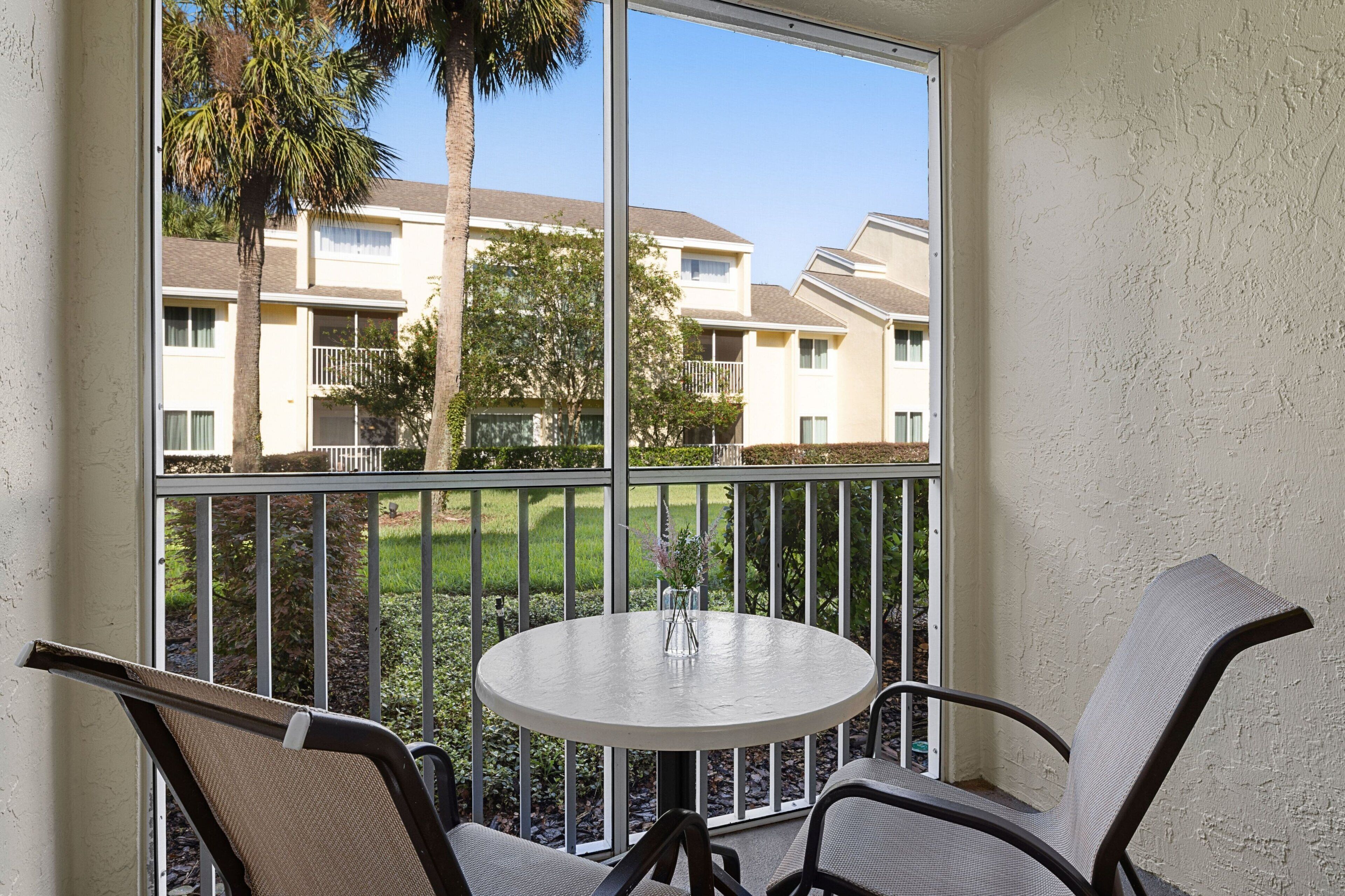 A balcony with a round table and two chairs, overlooking a residential area with apartment buildings and a lawn.