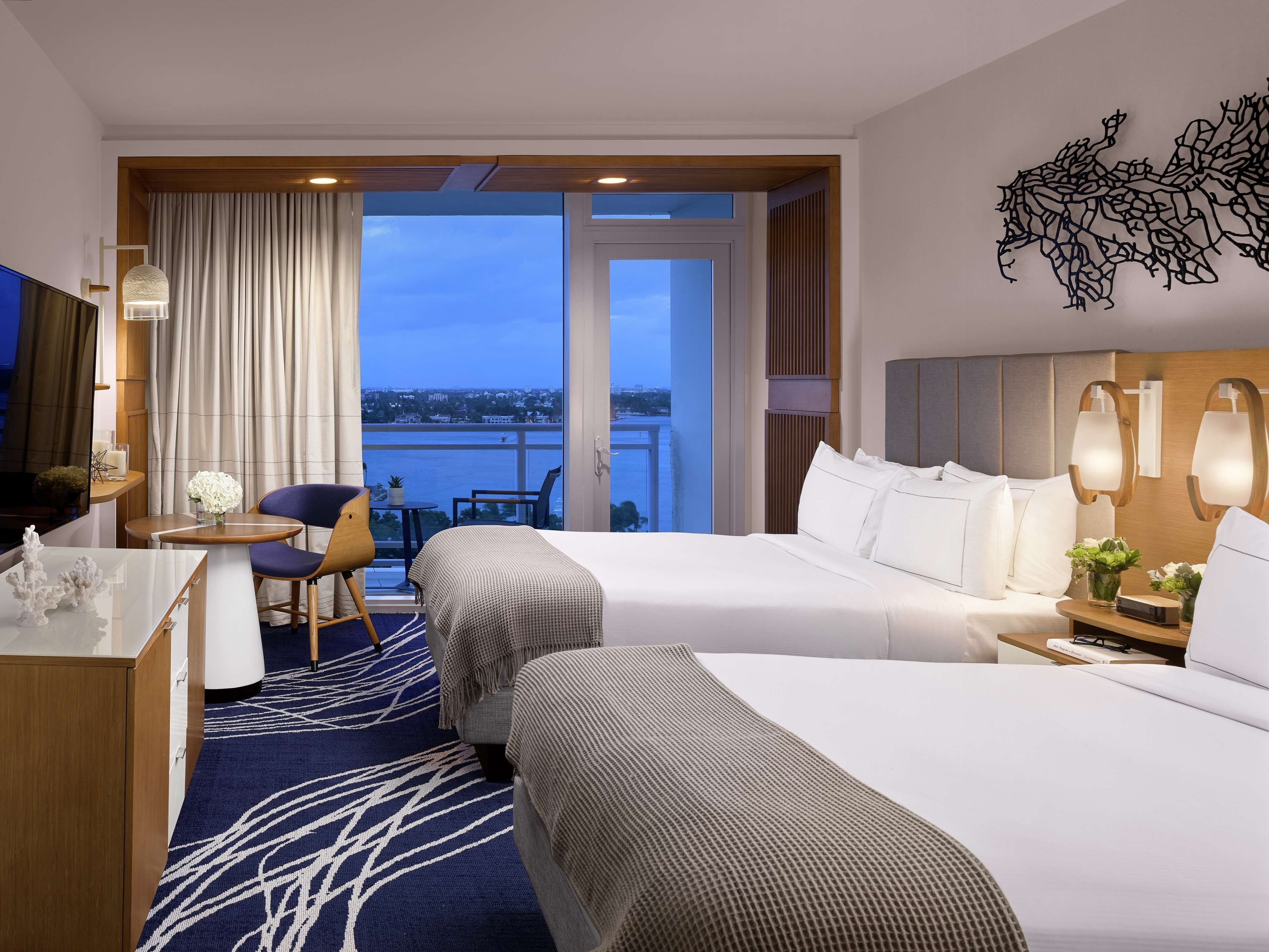 A hotel room with two beds, a desk, a chair, a small table, and a view of the city.