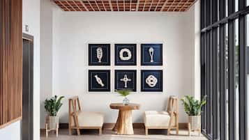 Un interior moderno con un entramado de techo de madera, una pared con seis diseños abstractos enmarcados, dos sillas, una mesa redonda de madera y plantas en macetas.