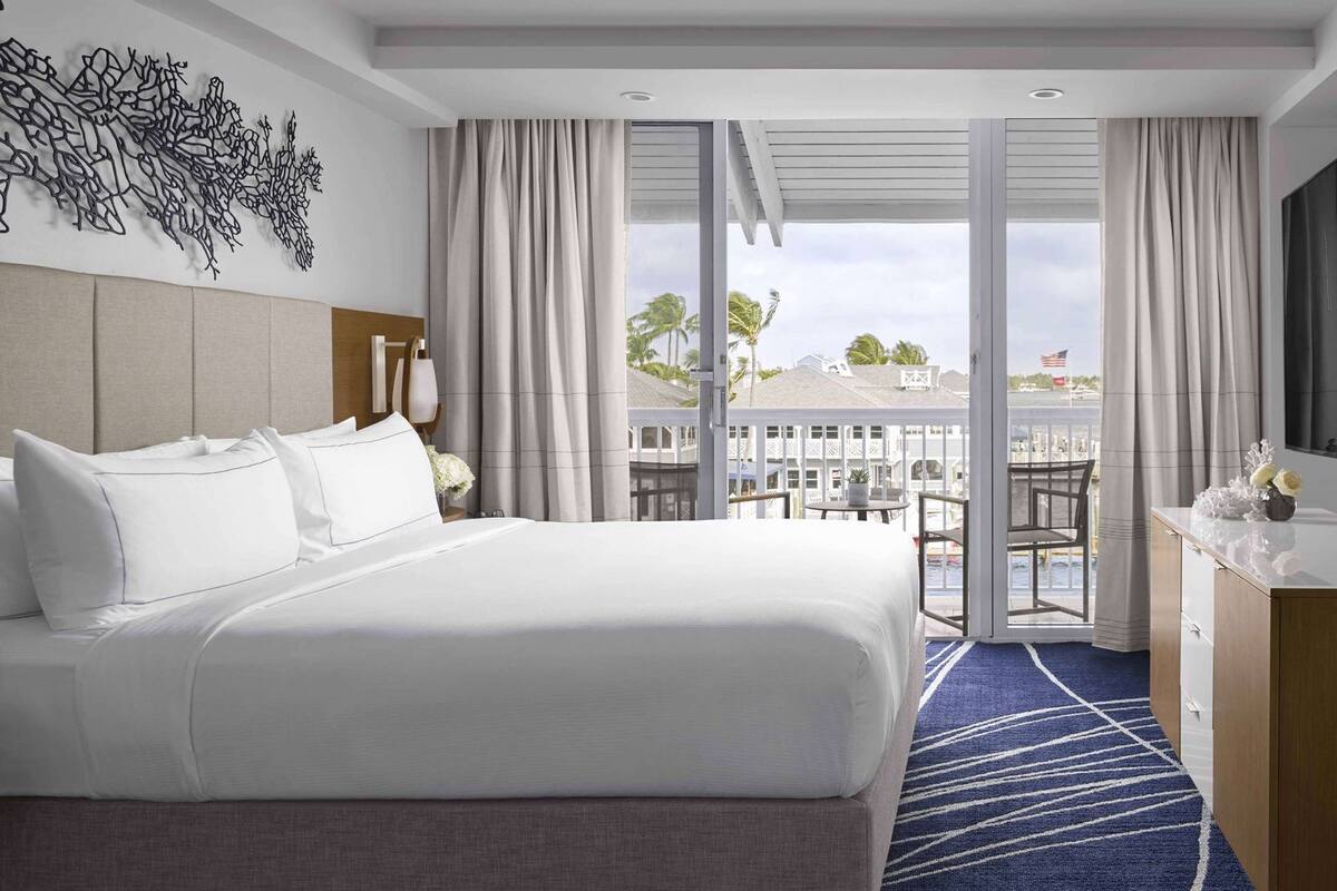 A hotel room with a large bed, a desk, a chair, a balcony with a view of the sea, and a decorative wall piece.