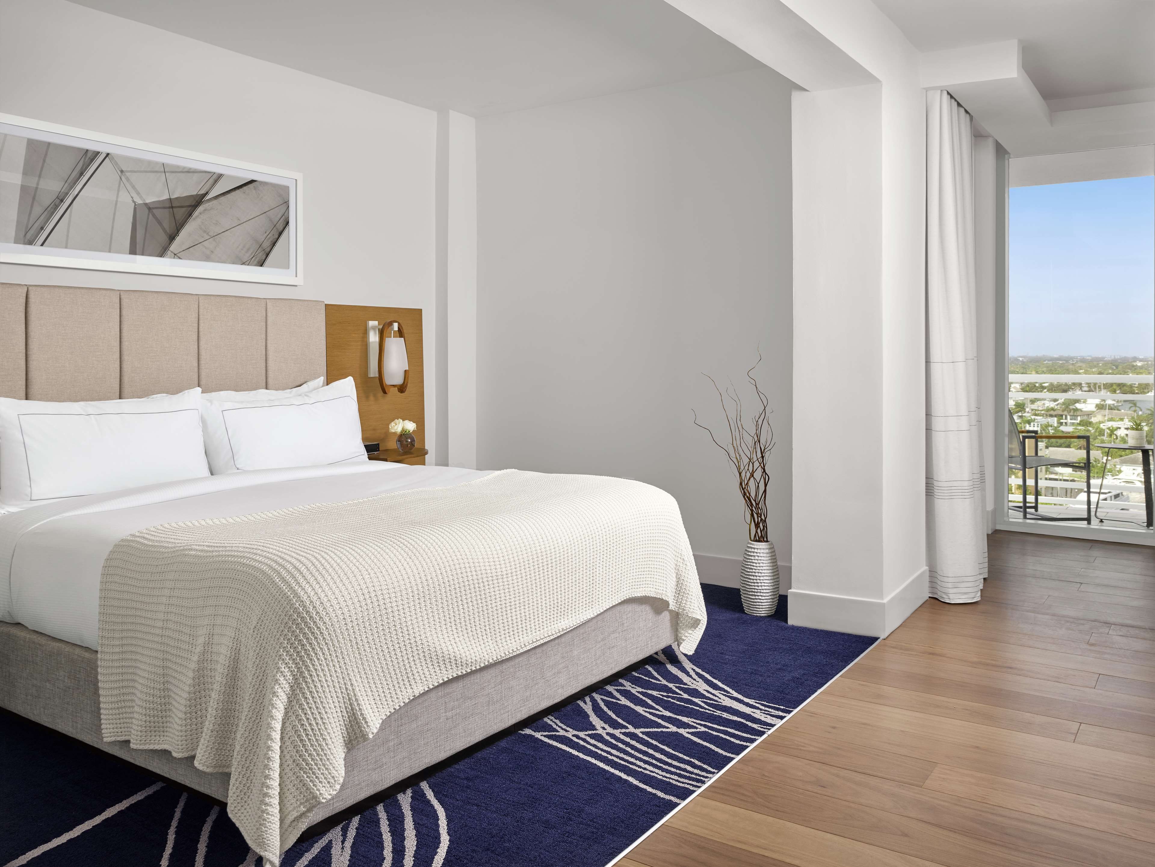 A hotel room with a bed, a nightstand, a vase with branches, a view of the city, and a balcony.
