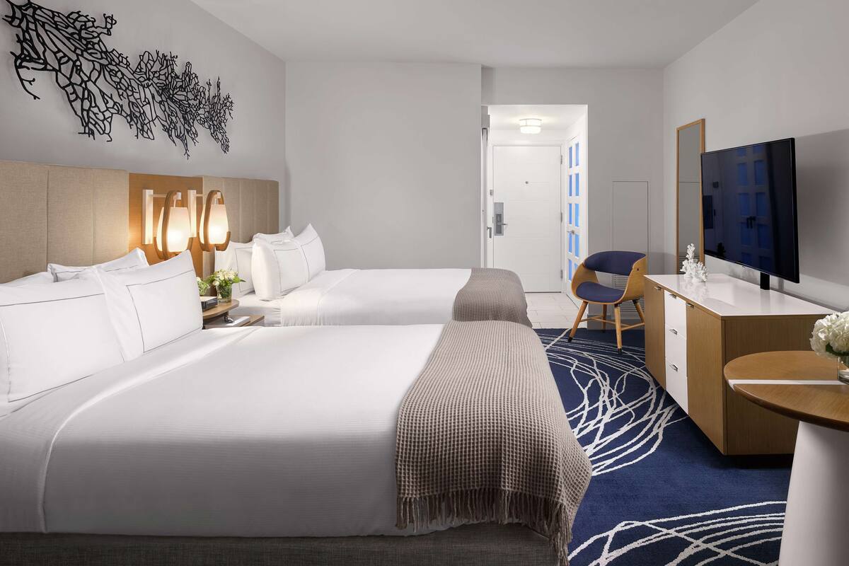 A hotel room with two beds, a desk with a TV, a chair, and a decorative wall piece.