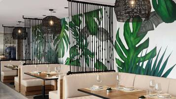 Un espace repas moderne avec des banquettes et des tables, agrémenté d’une fresque représentant des feuilles tropicales et de lampes noires suspendues.
