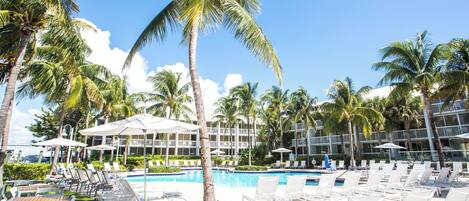A pool area with lounge chairs and umbrellas, surrounded by palm trees and a building with balconies.