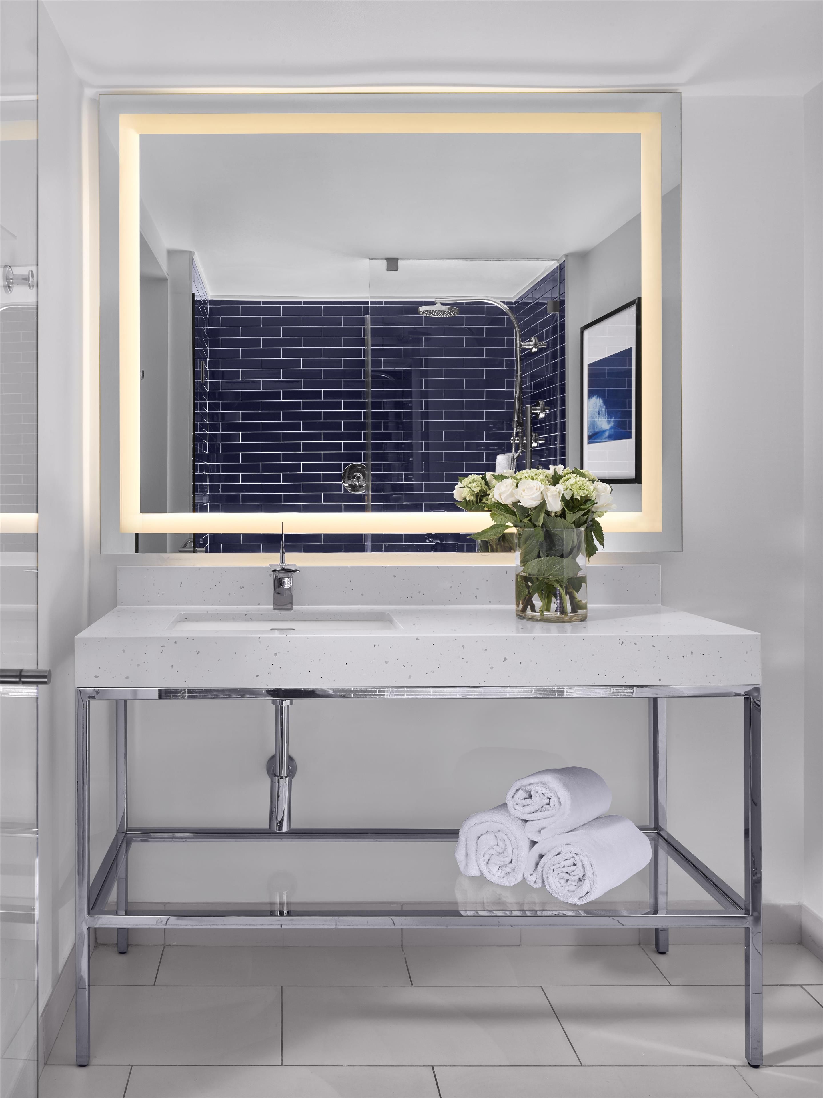 Un baño moderno con un lavamanos blanco, un espejo con marco iluminado, una ducha y un cuadro en la pared.