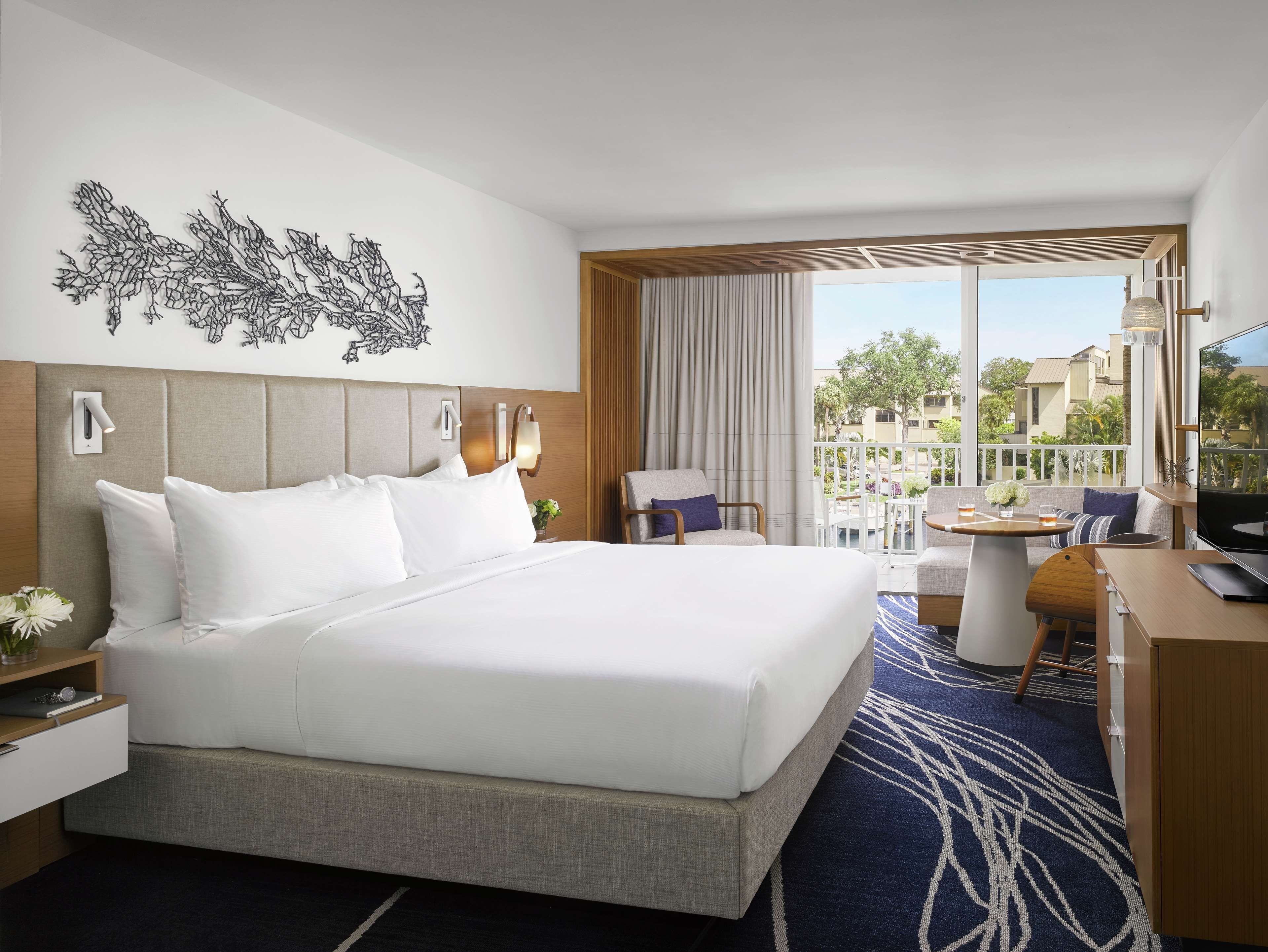 Habitación de hotel con una cama grande, un escritorio, una silla y un balcón con vistas a edificios y árboles.