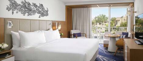 Habitación de hotel con una cama grande, un escritorio, una silla y un balcón con vistas a edificios y árboles.