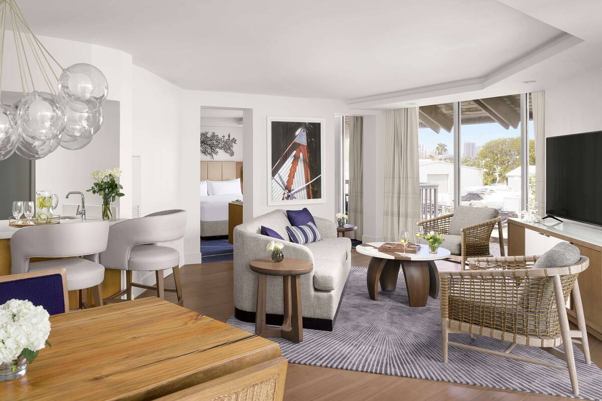 A modern hotel room with a sofa, chairs, a dining table, a TV, and a large window with a city view.