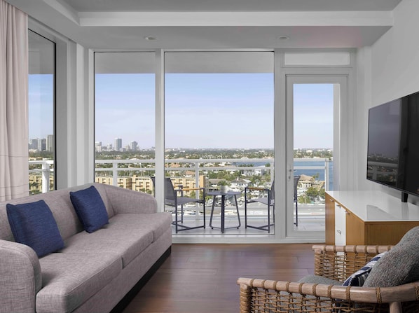 A modern living room with a sofa, a television, and a view of a cityscape through large windows.
