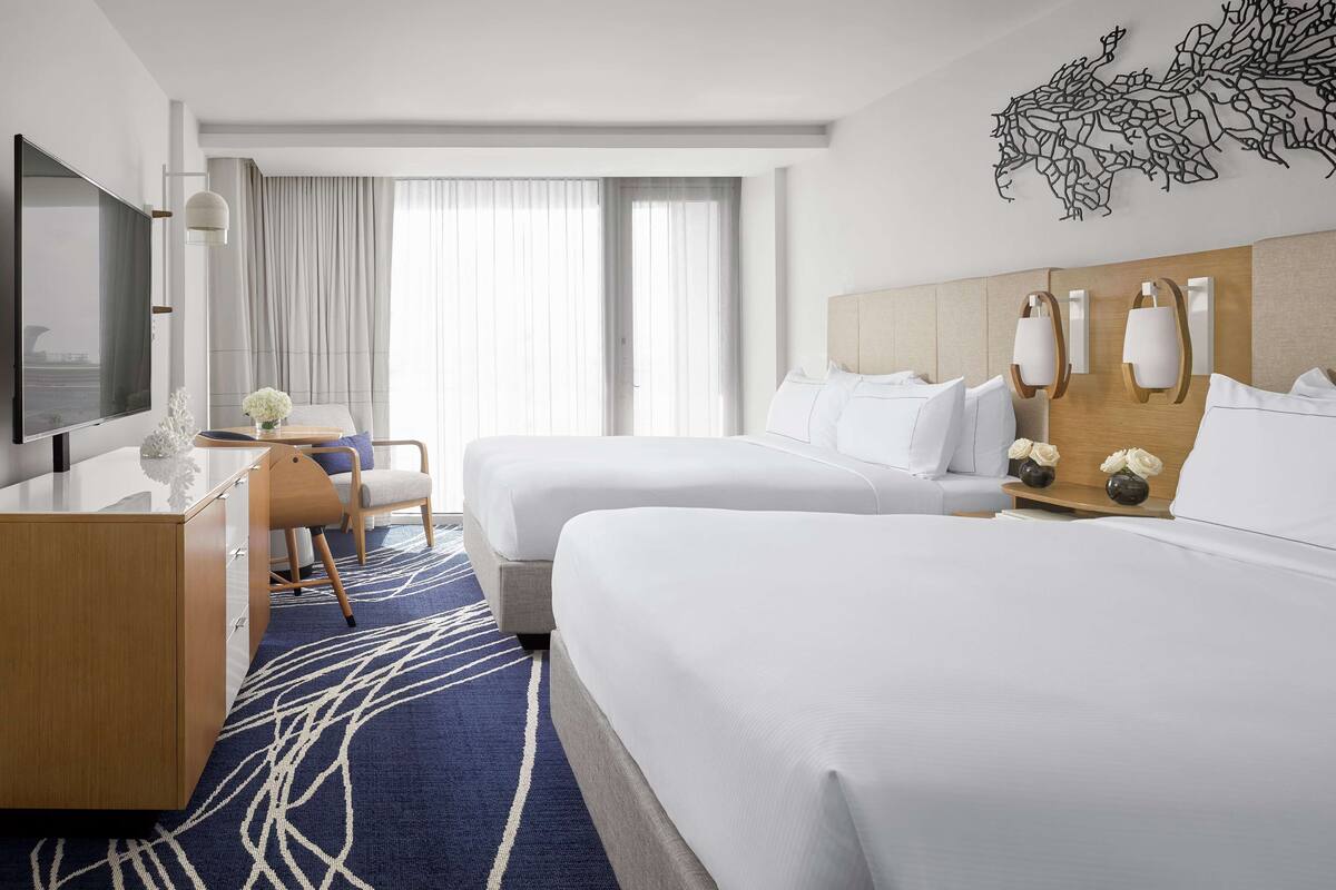 A hotel room with two beds, a desk, a TV, and a large window with curtains.