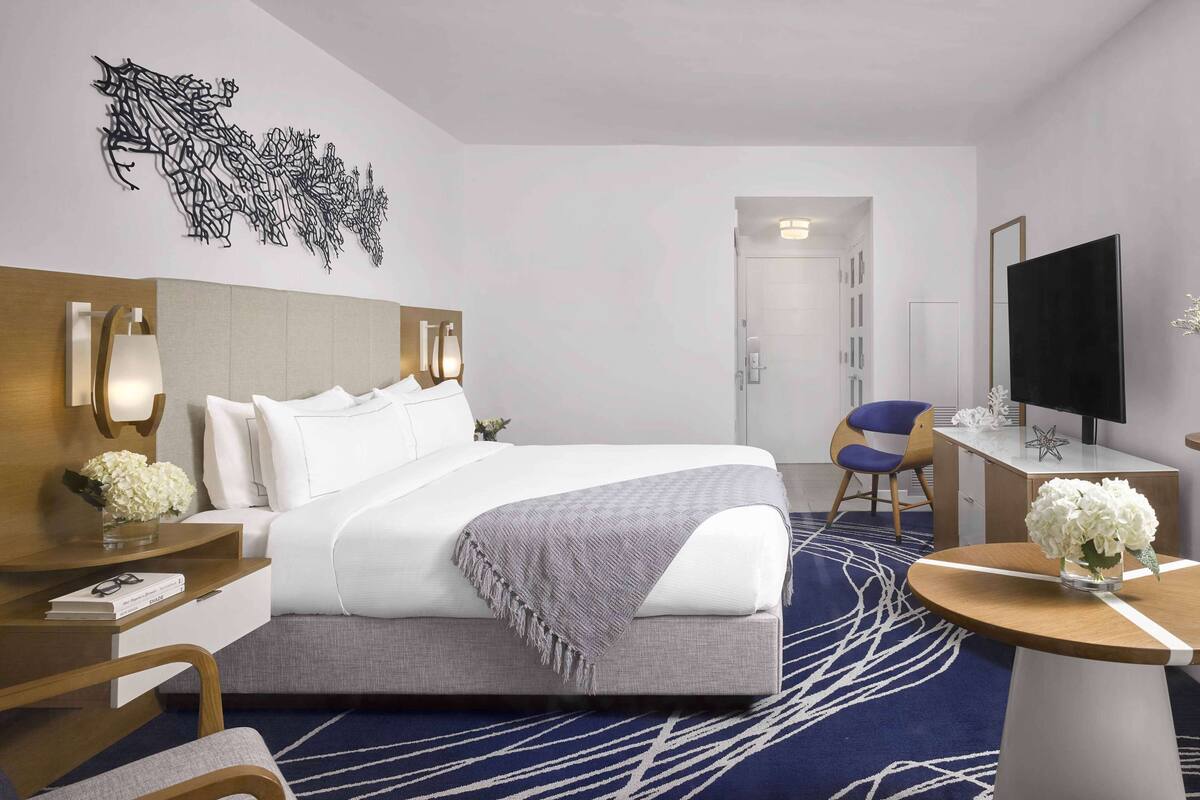 A modern hotel room with a large bed, a desk with a flat-screen TV, a chair, and a decorative wall piece.