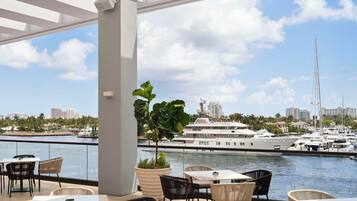 An outdoor seating area with tables and chairs, overlooking a marina with yachts and a city skyline.