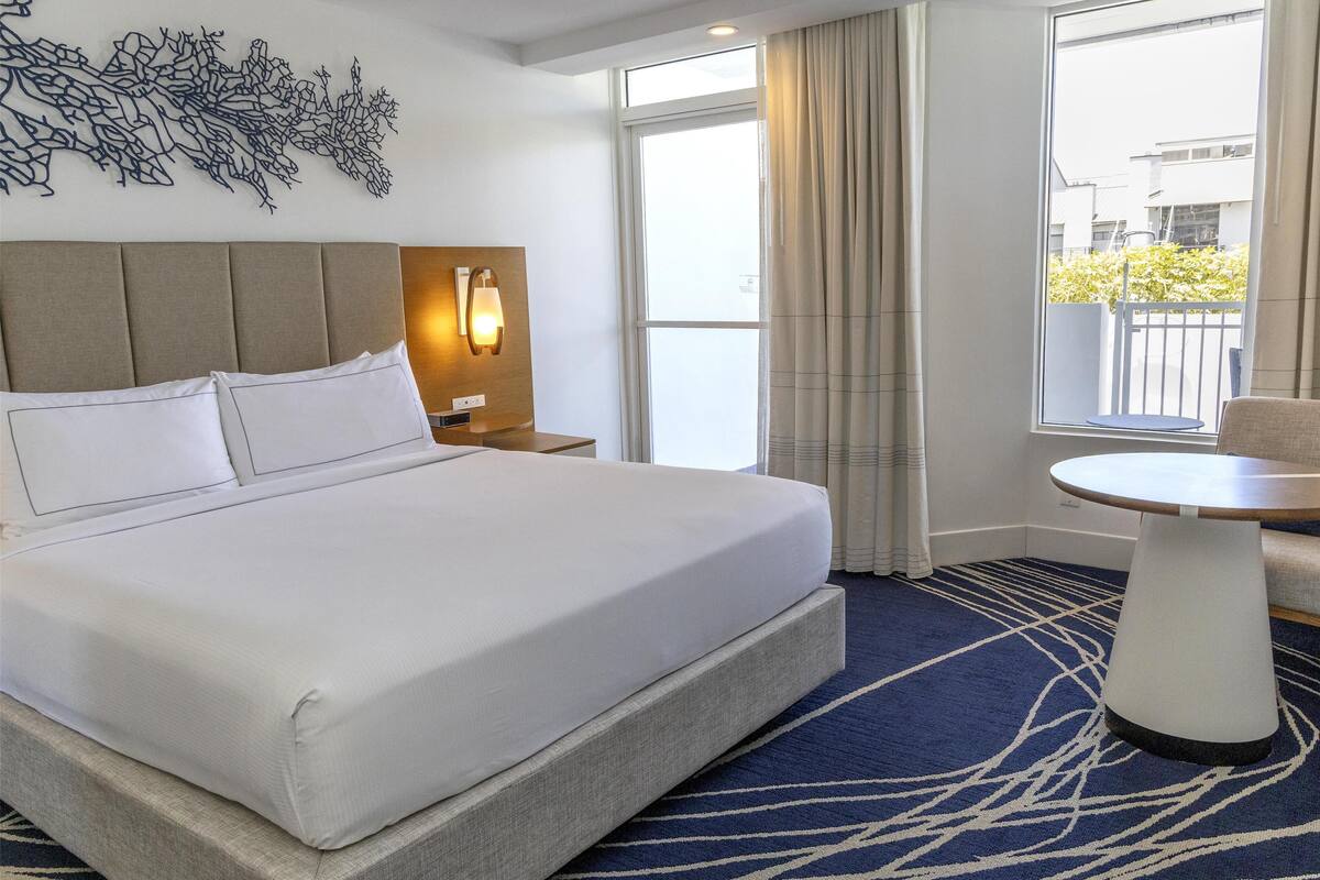 A hotel room with a large bed, a nightstand with a lamp, a small table, and a view of the outside through a window.