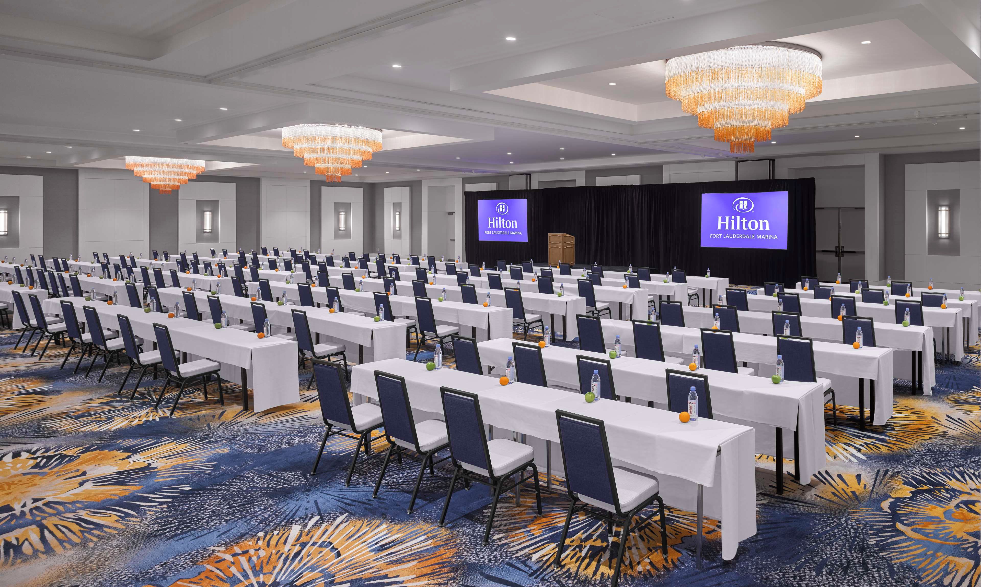 A large conference room with rows of tables and chairs, a stage with a screen, and chandeliers.