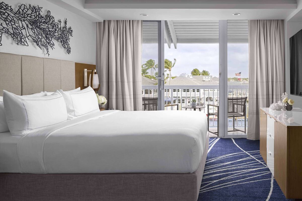 A hotel room with a large bed, a desk, a chair, a view of the ocean, and a balcony with a railing.