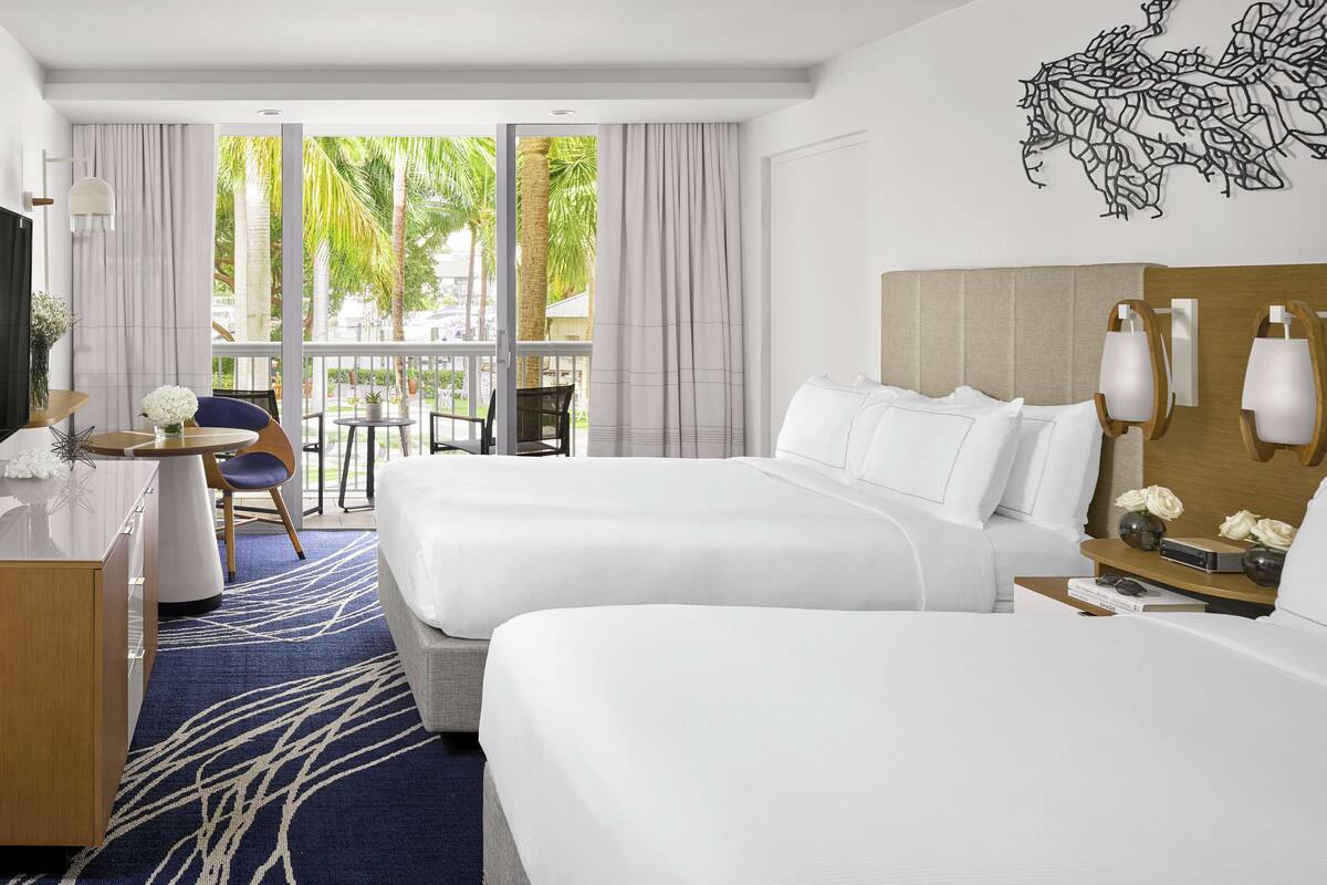 A hotel room with two beds, a desk with a chair, a TV, and a balcony with a view of palm trees.