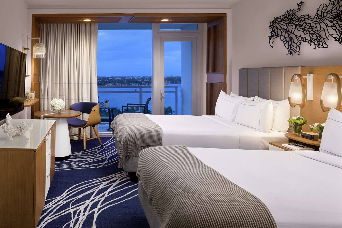 A hotel room with two beds, a desk, a chair, a small table with a vase of flowers, and a view of the cityscape through large windows.