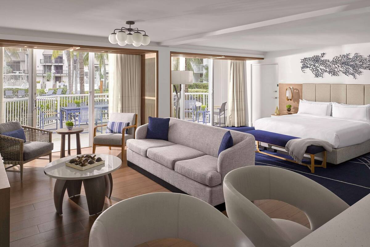 A hotel room with a bed, a sofa, a chair, a small table, and a balcony with outdoor furniture.