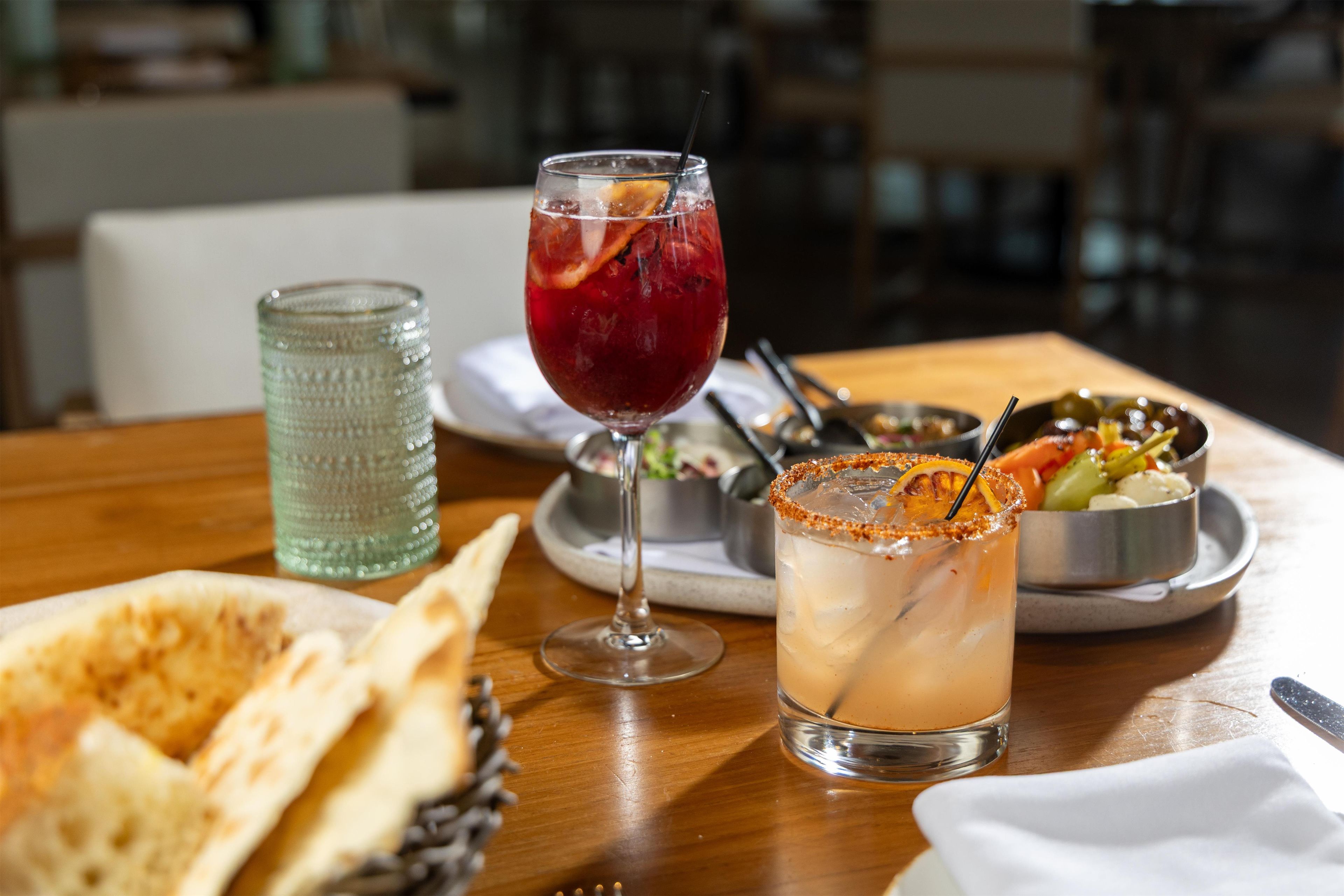 Una mesa de comedor con pan plano, un vaso de bebida roja con una rodaja de limón, una margarita con borde de sal y un vaso verde.