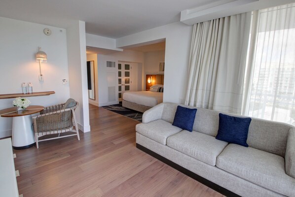 A modern hotel room with a sofa, a small table, a wicker chair, a bed, and a view of the city through large windows.